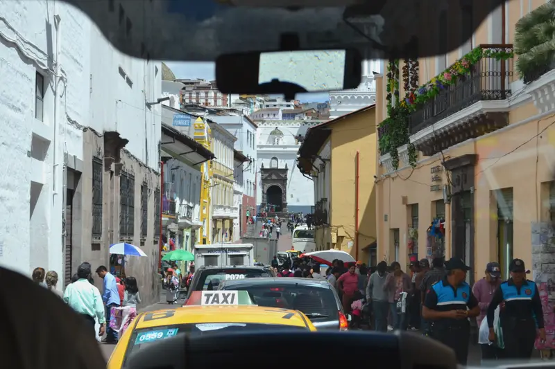 A narrow street in Quito's old town seen through a windscreen, taxis and pedestrians filling the road.