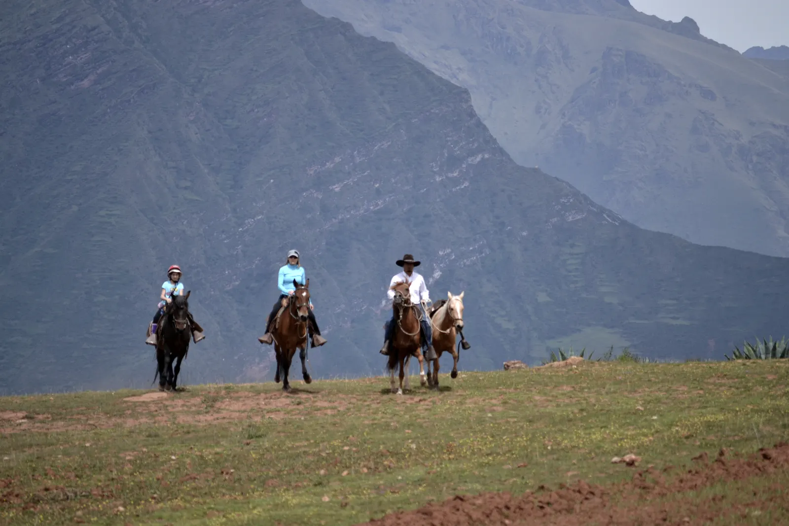 Three riders on a ridge above the Sacred Valley, the far wall of the valley rising behind them.