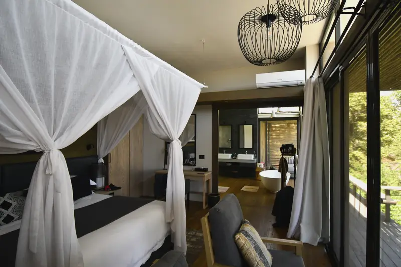 A suite interior with a four-poster bed, mosquito netting tied back, and a wall of glass onto the forest.