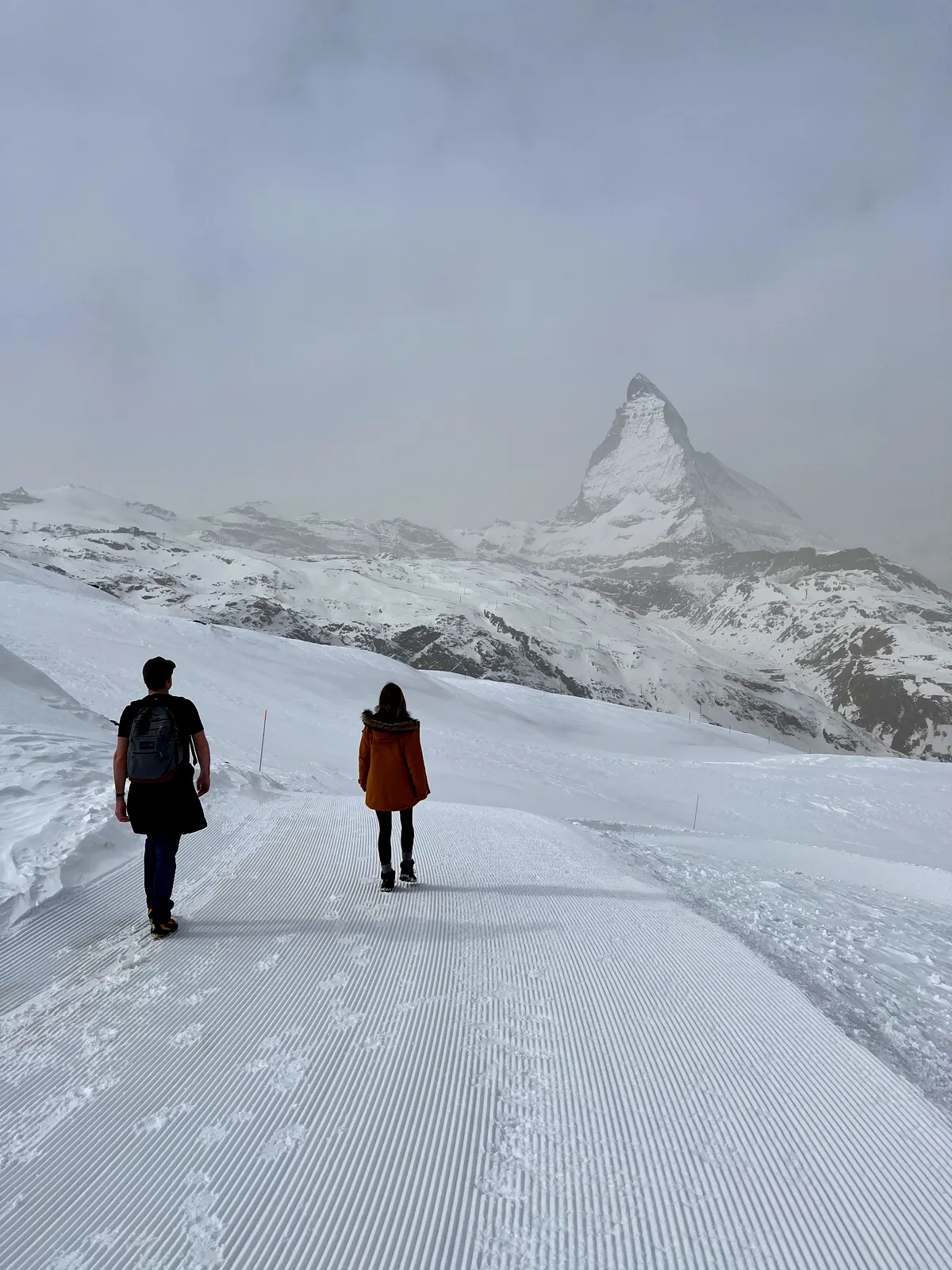 Two walkers on a groomed track above Zermatt, the Matterhorn standing at the end of it.