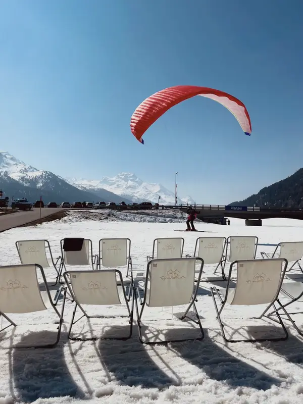 A paraglider coming in over a row of empty deck chairs on the snow.