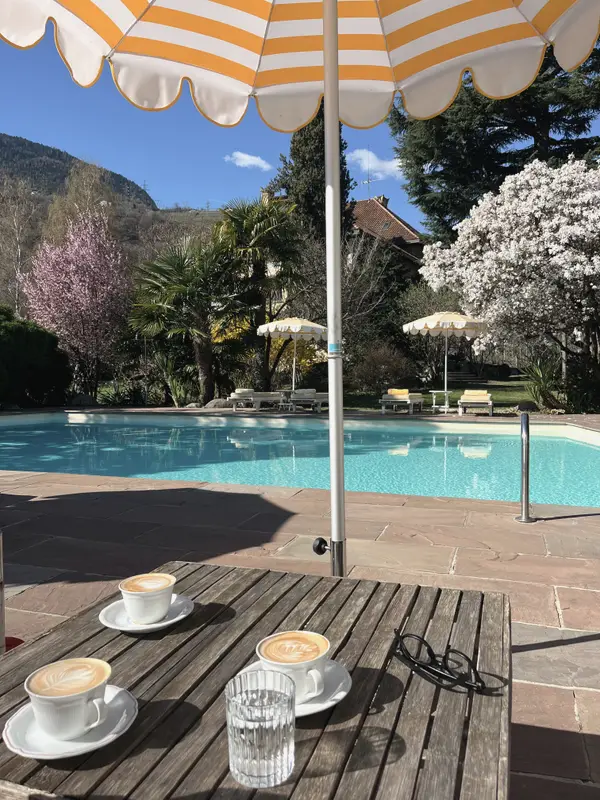 Two cappuccinos on a table by the pool, blossom and an orange parasol above.
